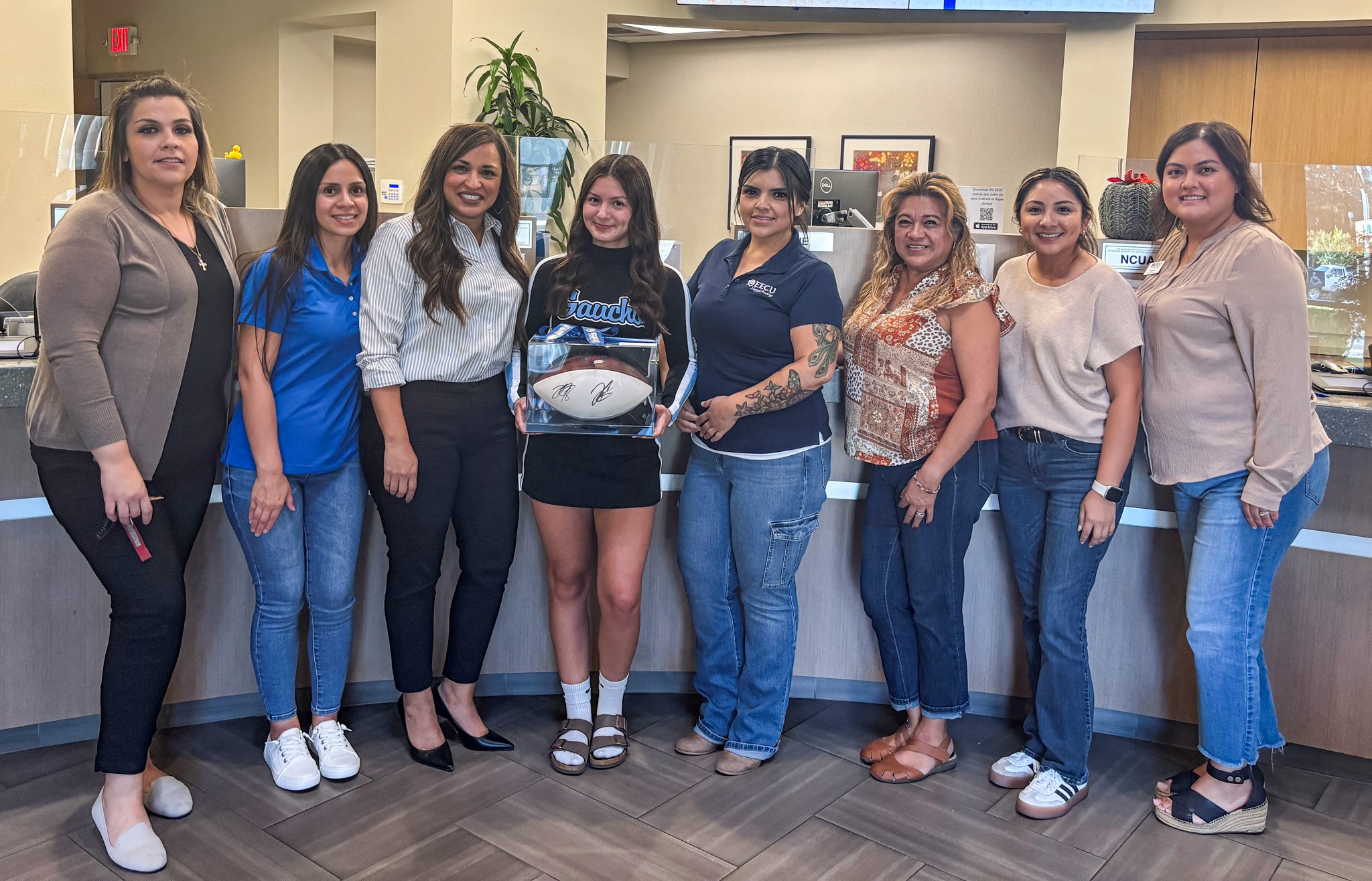 Seven EECU Employees inside a branch presenting a signed football to a El Capitan High School cheer leader